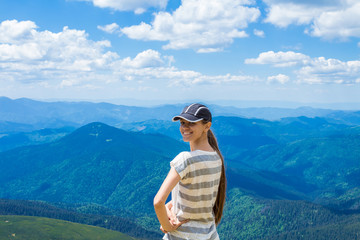 young tourist girl with long hair in a cap on top of the mountains, beautiful mountains and clouds on the background