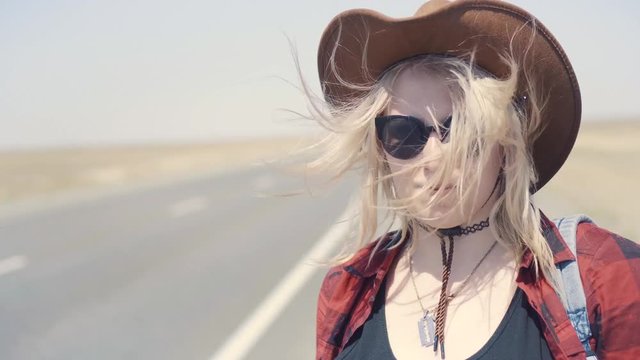 Young stylish girl in cowboy clothes doing hitch-hiking in desert, on the road. She carries backpack and hat. Pass trucks, delivery trucks. California or Nevada. Search yourself, escape from problems