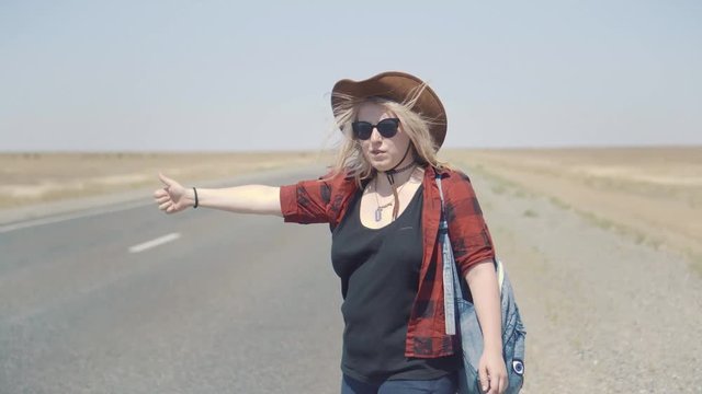 Young stylish girl in cowboy clothes doing hitch-hiking in desert, on the road. She carries backpack and hat. Pass trucks, delivery trucks. California or Nevada. Search yourself, escape from problems