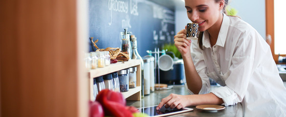 Young woman using tablet in kitchen at home and drinking coffee