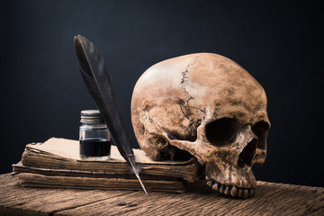 still life photography :  feather pen, inkwell and human skull on old book against art dark background