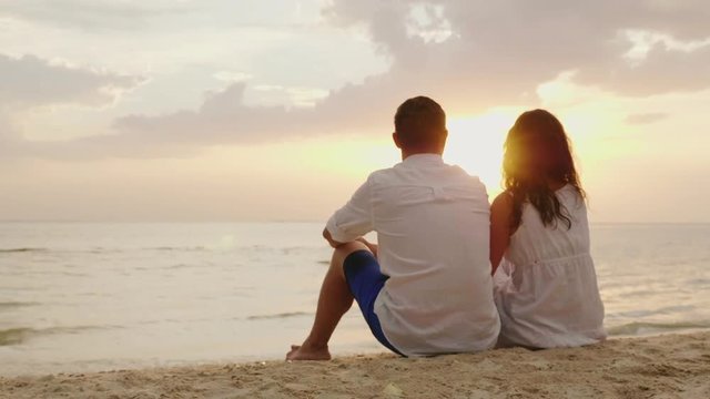 A man and a woman are sitting side by side on the sand on the beach. Together they look at the sunset over the sea