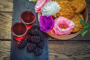 Homemade berry liqueur with fresh ripe blackberries, colorful eustoma flowers and sesame biscuits  in a wooden box and black stone tray on wooden background. Sweet alcohol drink.
