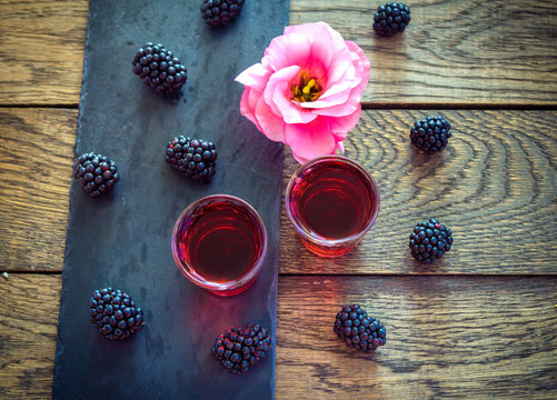 Homemade berry liqueur with fresh ripe blackberries, pink flower and black stone tray on wooden background. Sweet alcohol drink.