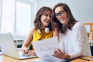 Couple of cheerful ladies working laughing while working together on laptop and documents in office