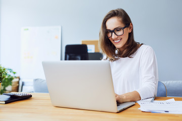 Attractive cheerful young businesswoman working on laptop and smiling while sitting at her desk in bright modern office