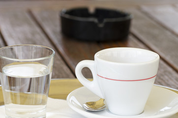 Close-up photo of a cup of coffee on a saucer with a spoon next to a glass of water and an ashtray
