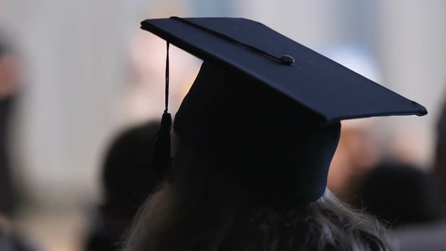 Brunette wearing hat and academic dress looking at stage and listening speech