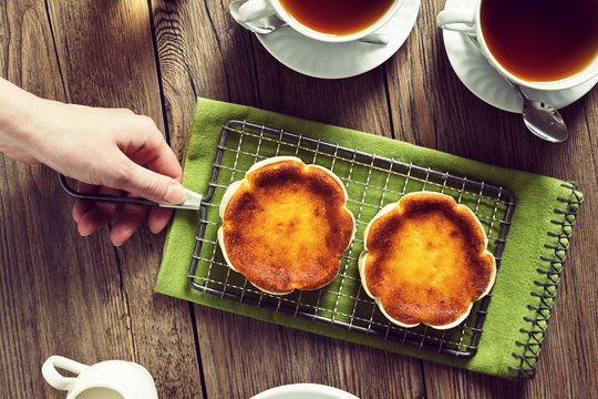 Woman's Hand Placing Queijadas or Queijadinhas, Mini Portuguese Baked Cheesecakes, Next to Cups of Tea on Table