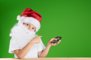Little Santa Claus looking into the camera. One hand shows on a green background, in the other hand holding a remote control. Remote is aimed at green background. Сhromakey. Close-up.