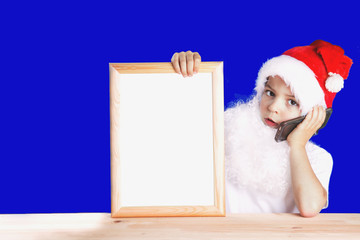 Little cute Santa Claus holding a blank picture frame with white background and talking on the phone. He looks into the camera. Blue background. Close-up.