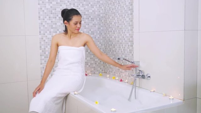 Video footage of a young woman with towel sitting on the bathtub while preparing hot water and playing soap bubble