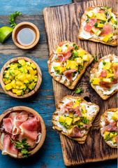 Toasts with cream cheese, ham jamon serrano and mango served on wooden board with red wine, blue wooden rustic background, top view