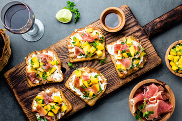 Toasts with cream cheese, ham jamon serrano and mango served on wooden board with red wine, gray slate background, top view