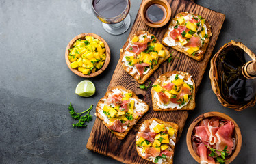 Toasts with mango, cream cheese, ham jamon serrano served on wooden board with red wine, gray slate background, top view. Copy space