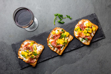 Toasts with cream cheese, ham jamon serrano and mango served on black slate board with red wine, top view, gray slate background