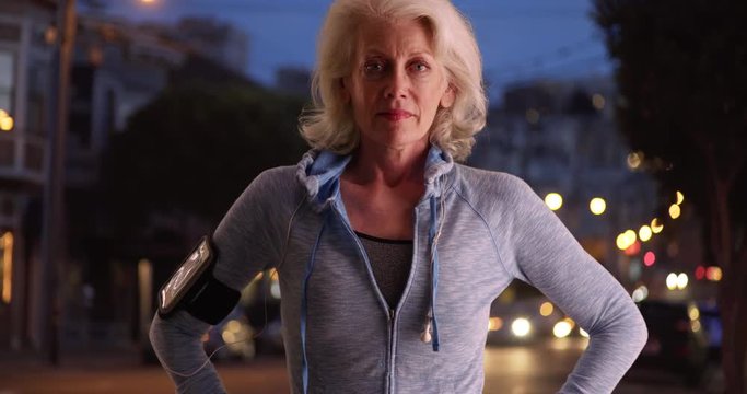 Portrait Of Female Jogger Looking At Camera Confidently In Urban Area In Evening. Athletic Older Woman In Her 50s Smiling At Camera Post Workout Outdoors At Night. 4k 