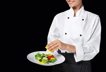 Young female chef dressing salad with lemon juice, on dark background