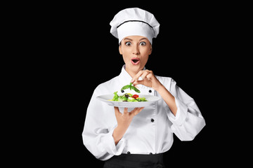Young female chef holding plate with salad on dark background