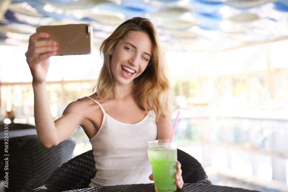 Poster Beautiful young woman with glass of lemonade taking selfie in cafe