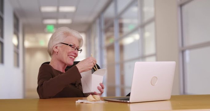 Mature Caucasian woman on lunch break at work eating Chinese takeout in office. Slow motion shot of businesswoman watching something on laptop while eating tasty noodles. 4k 