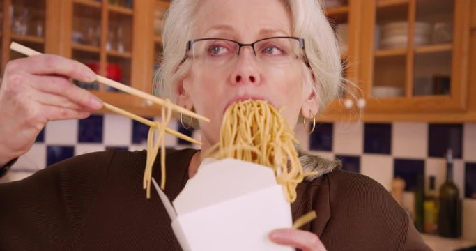 Slow motion of mature woman messily eating noodles out of takeaway box. Close up of Caucasian woman in her 50s eating Chinese takeout for dinner inside kitchen. 4k 