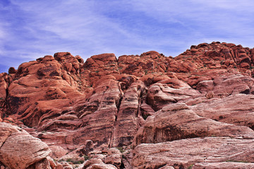 Looking upward at a cliff of jagged, craggy, red rocks with a blue, cloudy sky in the background. Red Rock, Nevada. There are small outcrops of dark green foliage and the rocks are cracked and eroded.