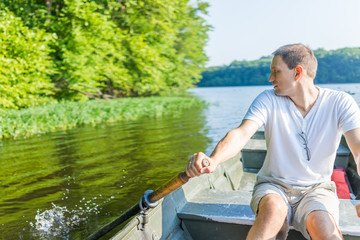 Fototapeta na wymiar Young man rowing boat on lake in Virginia during summer in white shirt looking back splashing water