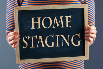 Female hands holding small black chalkboard in front of the body closeup with words Home Staging. On gray background with copy space