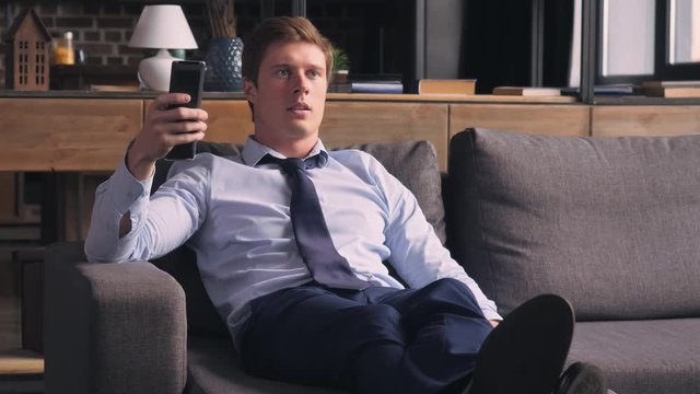 Young businessman in a bad mood at home. Caucasian handsome man resting in flat lying on sofa and watching tv changes channels. Male wearing in elegant clothes and tie.