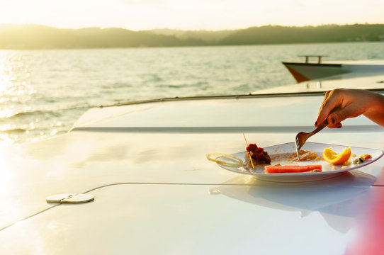 Hand With Fork Eating Food On The Deck Of A Yacht. A Beautiful Golden Lens Flare Adds To The Rich Affluent Theme Of The Shot