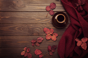 Knitted scarf of burgundy color with autumn leaves and a cup of coffee on a dark wooden background.
