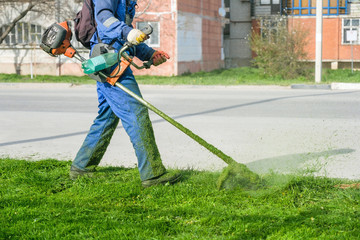 Man wearing overalls and rubber boots mowing green grass with gasoline lawn trimmer.