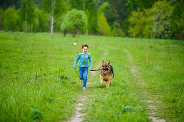 The little boy runs through the green meadow with his big dog of the breed German Shepherd