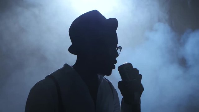 Man Young African American In Half Of The Turnover In The Smoke And White Light Approaching The Microphone Singing In A Recording Studio. Black Background. Silhouette. Close Up