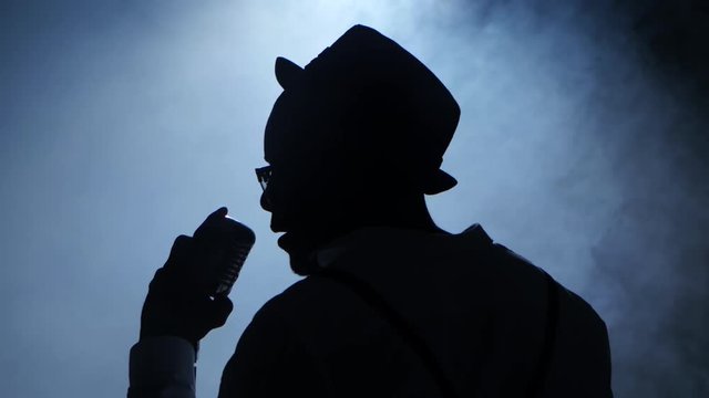 Singer the half of the turnover in the smoke and white light holding his microphone and gesticulating hands sings. Black background. Silhouette. Close up