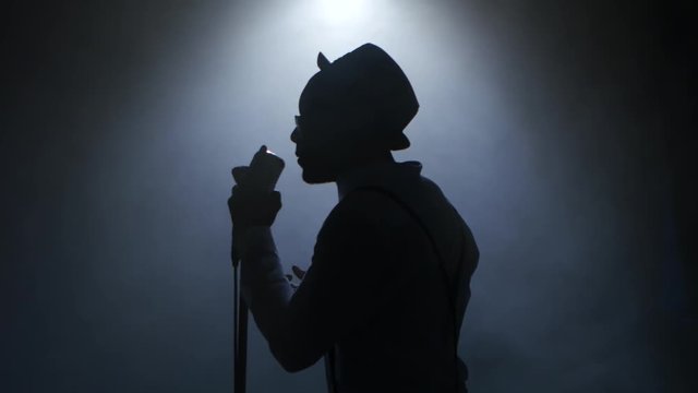 Man african american in half of the turnover in the smoke and white light approaching the retro microphone singing in a recording studio. Black background. Silhouette. Close up
