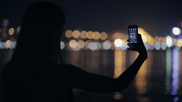 Woman photographing a night cityscape on smartphone standing near river