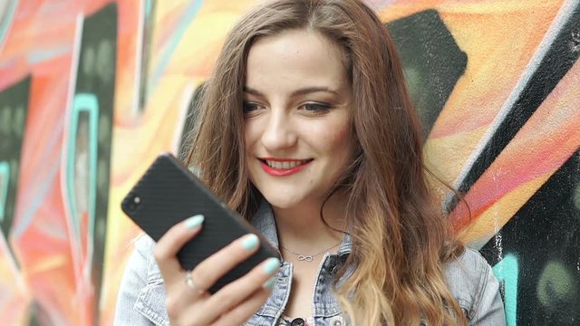 Pretty brunette leaning on the graffiti wall and doing selfies on smartphone, steadycam shot
