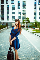 Young attractive woman with long legs with long hair in a blue elegant dress with a sword belt with a black bag in his hand, strolls in a European modern courtyard, a warm summer day