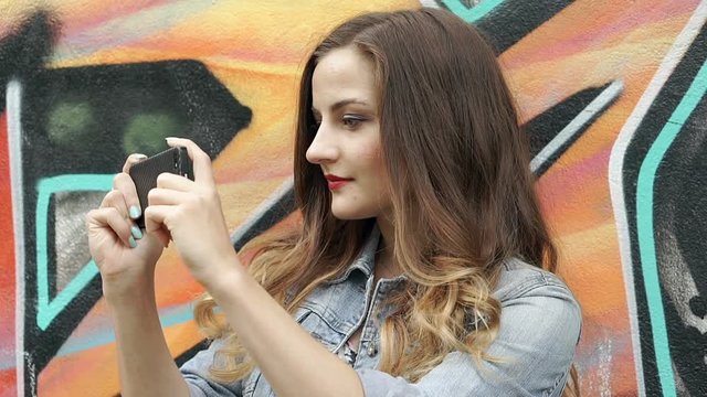 Pretty brunette standing next to the graffiti wall and doing photos on smartphone, steadycam shot

