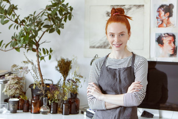 Lovely ginger female studying art in workshop, smiling gently in camera, standing crossed hands against beautiful pictures. Woman with red knot wearing apron going to creat something original