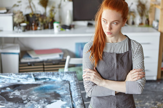 Beautiful red haired female wearing apron, keeping hands crossed, looking pensively aside while standing in workshop, resting for minute after creating masterpiece. Creative ginger young woman