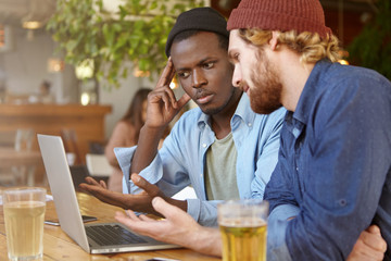 Picture of Afro American male using laptop computer during meeting with his Caucasian business partner at cafe for discussing business strategy and plans over a couple of beers, People and technology