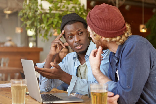 Interracial friendship concept. Two best friends wearing hats sitting at cafe table and talking, discussing plans, sharing news, drinking beer and watching football match on generic latop computer