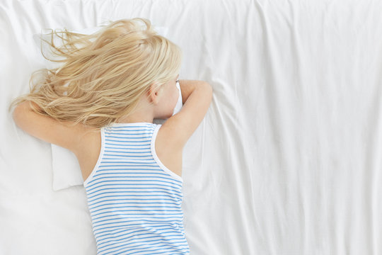 Back View Of Adorable Blonde Girl Wearing Striped T-shirt, Having Healthy Sleep, Lying On Stomach On White Pillow, Dreaming About Something. Restful Carefree Small Kid Sleeping In Bed After School