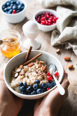 Oatmeal porridge bowl with superfoods in hands. Hands holding bowl of oat porridge with blueberries, almonds, linseed, cinnamon and currants. Selective focus