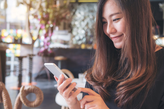 Closeup image of a smiley beautiful Asian woman using and looking at smart phone in modern cafe