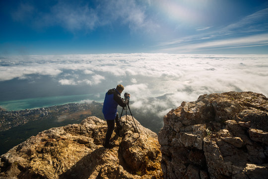 Man standing with a tripod and camera on a high mountain peak above clouds, city and sea. Professional photographer adjusting dslr settings on rocky summit.