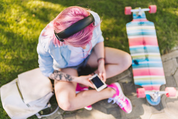 Top view of European beautiful woman with pink hair in headphones listening to music from smartphone sitting in park on green grass background in a blue shirt. 